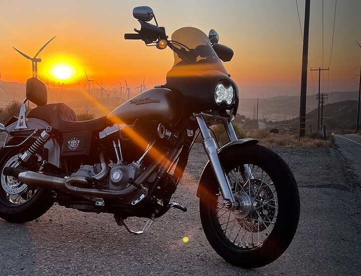 Motorcycle at sunrise with a SlimLite 6” LED headlight shining on an open highway, wind turbines in the background.