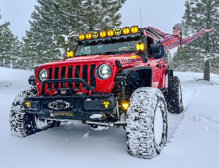 Custom red Jeep Rubicon in the snow with LED lights and smiley covers, as a woman leans out, enjoying her off-road adventure.